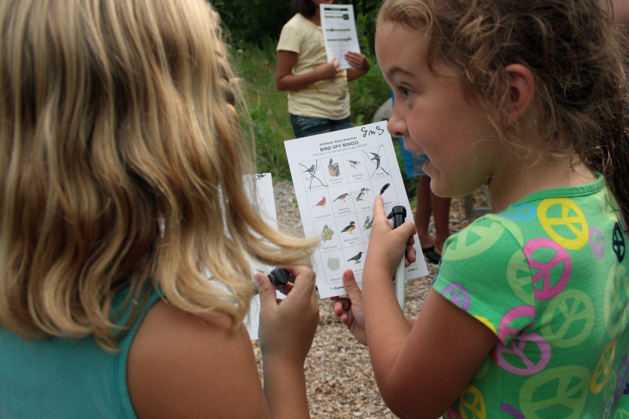 Two girls holding Bird Bingo cards and a marker. One is pointing a finger at the card.